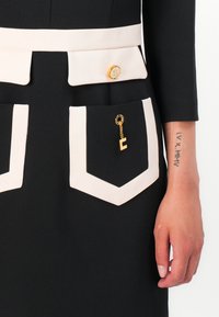 Black dress featuring white pocket accents, a gold button, and a small gold charm with an "E" hanging from the pocket chain.