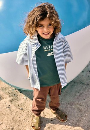 Bambino piccolo con capelli ricci sorridente, indossa una maglietta verde "Mexico", una camicia a maniche corte a righe, pantaloni marroni e sandali, in piedi all'aperto sulla sabbia.