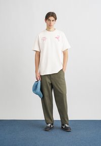 Ανοιχτό ροζ βαμβακερό μπλουζάκι με γραφικά σχέδια, συνδυασμένο με λαδί φαρδείς παντελόνες. Κρατάει ένα ανοιχτό μπλε καπέλο. Μαύρα παπούτσια.