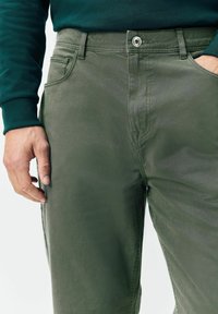 Mees, kes kannab tumehaldeid pükse ja tumeteal särki pikkade varrukatega, mille ühe käe on ta pannud tasku ja teine on lõdvestunult külje ääres.