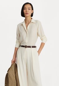 Light beige button-up shirt with a collar, paired with high-waisted cream trousers and a brown leather belt. Structured brown herringbone blazer.