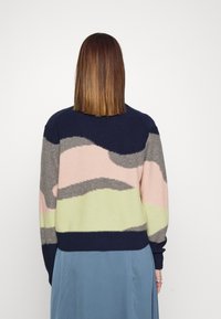 Femme aux cheveux raides mi-longs, portant un pull multicolore à motif abstrait et une jupe bleue, vue de dos sur un fond blanc uni.