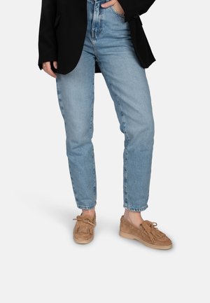 Helle blaue Denim-Jeans mit entspannter Passform und fransenbesäumtem Saum, kombiniert mit tanen Wildlederschuhen mit Fransen- und Schnürdetails. Schwarzer Blazer trägt.