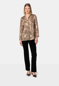Blusa marrone con stampa serpente, scollo a V e maniche lunghe, abbinata a pantaloni in velluto nero e décolleté marroni a punta.