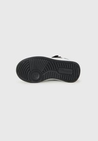 Semelle d'une sneaker noir et blanc présentant des motifs de traction texturés et un logo circulaire sur le talon, présentée sur un fond clair.