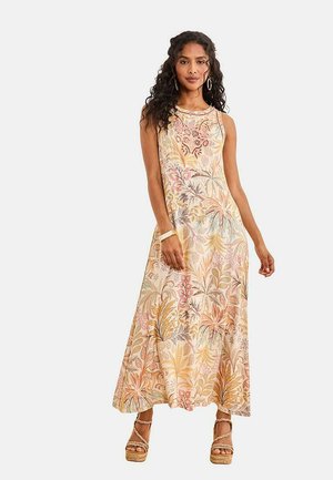 Mujer de pie con un vestido floral sin mangas hasta el suelo con un patrón de hojas en tonos pastel y sandalias de cuña, posando con una mano hacia abajo.