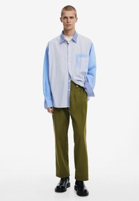 Gestreiftes Hemd in Blau und Weiß mit einer Brusttasche, kombiniert mit olivgrünen, plissierten Hosen und schwarzen Stiefeln. Glatte Textur.