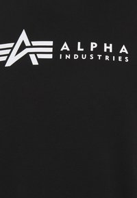 Juodas marškinėliai su balta spausdinta grafika, kurioje pavaizduota "ALPHA INDUSTRIES" logotipas su kampuotais akcentais, pateiktas drąsiais, modernaus stiliaus šriftais.
