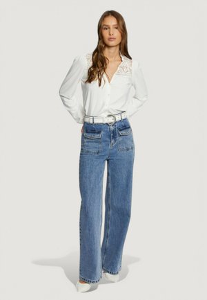 Camicia bianca con bottoni e dettagli in pizzo sulle spalle, abbinata a jeans blu a vita alta e una cintura bianca.