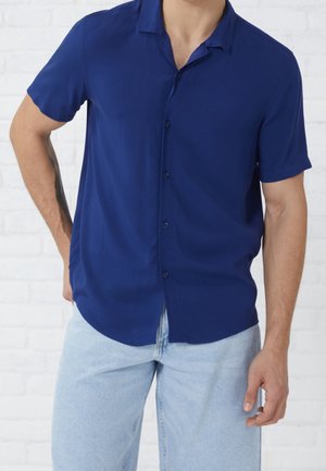 Mężczyzna w granatowej koszuli na krótki rękaw z guzikami i jasnoniebieskich dżinsach, stojący z jedną ręką w kieszeni na tle białej ceglanej ściany.