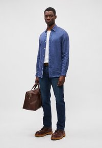 Μπλε λινό πουκάμισο, λευκό μπλουζάκι από κάτω, σκούρο μπλε τζιν, καφέ δερμάτινα παπούτσια με χοντρές σόλες και καφέ δερμάτινη τσάντα.