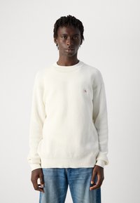 Persoon in een witte, gestructureerde gebreide trui met een klein CK-logo patch, gecombineerd met blauwe jeans, staand tegen een effen lichte achtergrond.