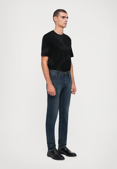 T-shirt preta de manga curta com padrão, jeans ajustados azuis e sapatos de pele pretos. A t-shirt apresenta um design texturizado. Modelo em pé.