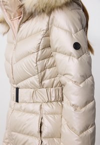 Béžový prošívaný zimní kabát s kapucí lemovanou kožešinou, páskem v pase a černým kruhovým logem na levém rukávu.