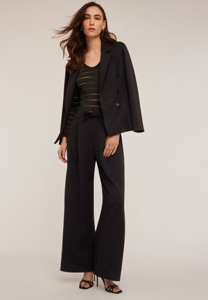 Schwarzer Wollmischung-Blazer mit doppelter Knopfleiste, kombiniert mit einem schwarz gestreiften Langarmoberteil und weiten schwarzen Hosen.