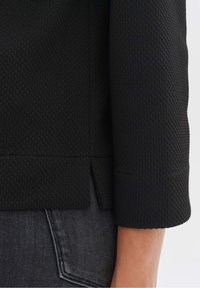 Manica di giacca nera con texture e un sottile motivo a nido d'ape, caratterizzata da un polsino dritto e un leggero spacco all'orlo, abbinata a jeans scuri.