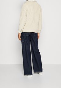 Chaqueta de pana beige con un acabado texturizado y cuello de pelo, combinada con pantalones cargo oscuros de mezclilla de pierna ancha con bolsillos laterales.