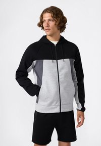Mann in schwarz, grau und hellgrauem Reißverschluss-Hoodie mit Kapuze und schwarzen Shorts, der mit einer Hand in der Tasche steht und nach links schaut.