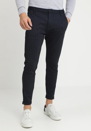 Homme portant un pantalon bleu marine à rayures fines, coupe ajustée avec ourlets retroussés, associé à des baskets blanches et un haut à manches longues gris clair.