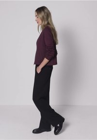 Weinrote Reißverschlussjacke mit Seitentaschen, kombiniert mit locker sitzenden schwarzen Hosen und schwarzen Schuhen, gezeigt an einem seitlich stehenden Model.