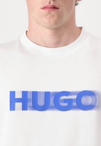 Hvid bomulds t-shirt med et blåt grafisk tryk af ordet "HUGO" på brystet. Rund halsudskæring og blød tekstur.