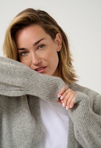 Grauer gestrickter Cardigan mit gerippten Bündchen, weicher Textur, lässiger Schnitt, über einem weißen Hemd getragen. Hände berühren sanft die Bündchen.