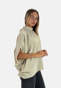 Top beige oversize con collo alto, maniche corte e tessuto testurizzato. Indossato con pantaloni grigio scuro, esposto su uno sfondo neutro.