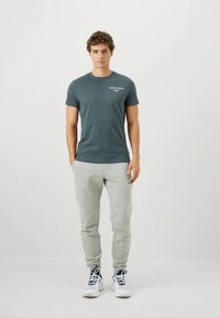 T-shirt cinzenta de algodão com mangas curtas, com um pequeno logótipo branco, combinada com calças de treino cinza claro e sapatilhas brancas com detalhes azuis.