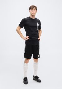 Μαύρο αθλητικό μπλουζάκι με γραφικό σχέδιο, σε συνδυασμό με μαύρο σορτς με λευκές ρίγες, λευκές κάλτσες μέχρι το γόνατο και μαύρα παπούτσια ποδοσφαίρου με πράσινες λεπτομέρειες.