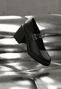 Μαύρο παπούτσι Mary Jane με τετράγωνο τακούνι, διπλές λουράκια και ασημένιες αγκράφες πάνω σε ανακλαστική ασημένια επιφάνεια με μαξιλαράκι.