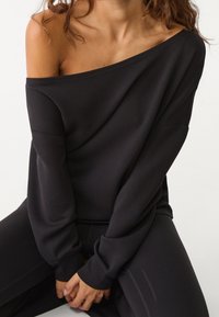 Sudadera negra de hombros descubiertos hecha de tela suave, con mangas largas y un corte holgado, combinada con leggings oscuros.