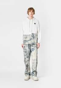 Sudadera blanca con un logo, combinada con pantalones de denim en tonos azul y gris con estampado. El modelo lleva unas zapatillas con suela gruesa blanca.