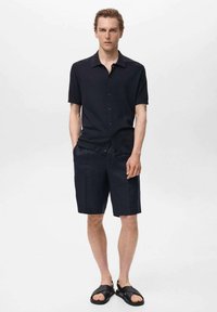 Camicia corta a maniche corte di colore scuro, in tessuto testurizzato, abbinata a pantaloni corti blu navy sartoriali e sandali neri, su uno sfondo neutro.