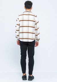 Homme de dos portant une chemise à carreaux blanche et marron, un pantalon noir et des baskets noires sur fond blanc uni.