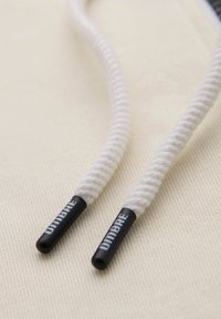 Gros plan des bouts de lacets blancs torsadés avec embouts en plastique noirs estampillés "DIMBRE" sur un fond de tissu beige.