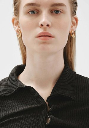 Se llevan pendientes de aro de oro con un diseño liso y curvado. El modelo viste una camisa negra texturizada con cuello y costillas verticales.
