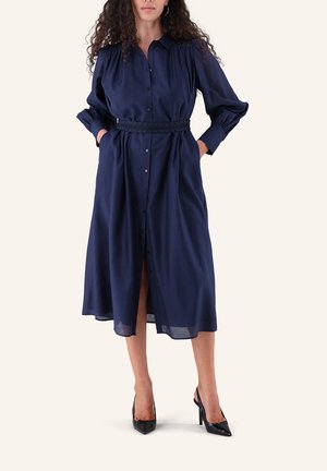 Frau trägt ein marineblaues Midi-Hemdblusenkleid mit langen Ärmeln, einem taillierten Gürtel und schwarzen spitzen High Heels.