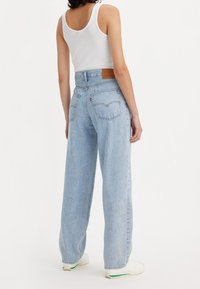 Personne portant un jean large taille haute bleu clair, un débardeur blanc et des baskets blanches, debout de dos sur un fond uni.