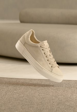 Baskets casual beige avec lacets et semelle blanche épaisse, photographiées en plein air au-dessus d’un tapis texturé et d’un canapé beige en arrière-plan.