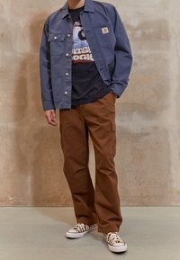 Person, der eine blaue Knopfjacke, ein schwarzes T-Shirt mit Grafik, braune Cargo-Hosen und schwarz-weiße Sneaker mit Muster trägt, steht vor einer beigen Wand.