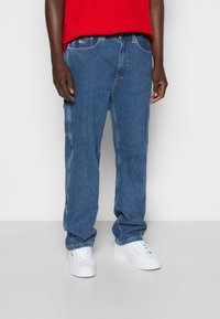 Jeans in denim blu con vestibilità rilassata, caratterizzati da molteplici tasche e un classico design a cinque tasche. Indossati con sneakers bianche.