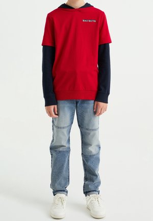Garçon portant un t-shirt rouge à manches courtes superposé à un t-shirt à manches longues bleu marine, un jean bleu clair avec des coutures au niveau des genoux, et des baskets blanches, debout.