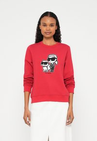 Nő piros pulóvert visel, amelyen egy rajzfilmfigura szerepel: egy ember és egy macska, mindketten fekete napszemüveget viselnek.