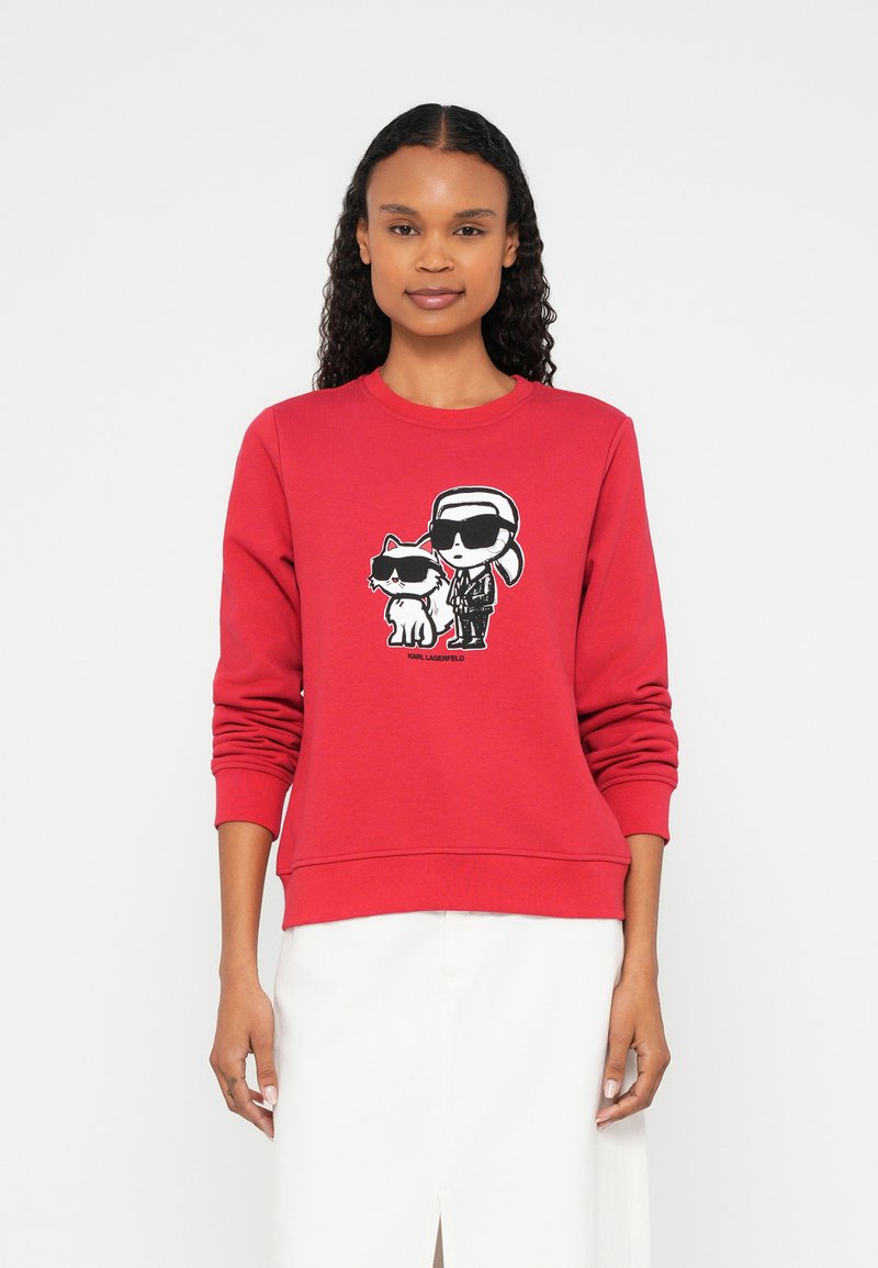 Nő piros pulóvert visel, amelyen egy rajzfilmfigura szerepel: egy ember és egy macska, mindketten fekete napszemüveget viselnek.