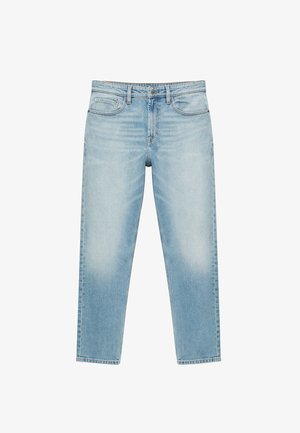 Person med mørk hud iført en hvid kortærmet skjorte og lyseblå jeans, med armene krydset over brystet.
