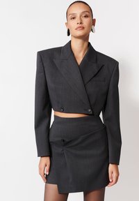 Kobieta z włosami zebranymi w kucyk, ubrana w ciemnoszary, krótki żakiet z szerokimi ramionami oraz dopasowaną, wysoką asymetryczną spódnicę, pozująca na tle białym.