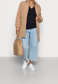 Beige vadderad lång kappa över en marinblå tröja, parat med ljusblå vida jeans och vita sneakers. Bär en beige dragkedjesäck.