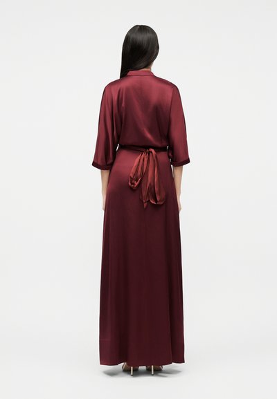 Γυναίκα με μακριά σκούρα μαλλιά, γυρισμένη από την πλάτη, φοράει μακρύ, μεταξένιο μπορντό φόρεμα με μανίκια τρία τέταρτα και ζώνη στη μέση δεμένη σε φιόγκο.