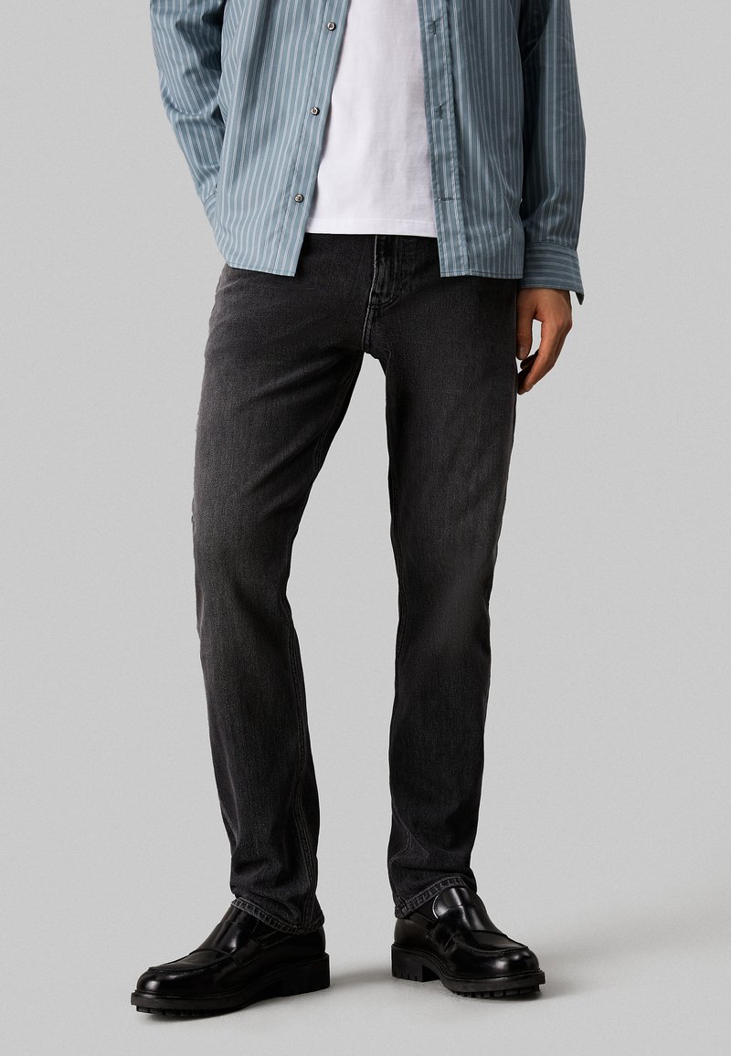 Pantalones vaqueros de mezclilla gris oscuro con un corte recto, combinados con una camisa de manga larga a rayas azul clara y zapatos negros. Sin patrones visibles en los pantalones.