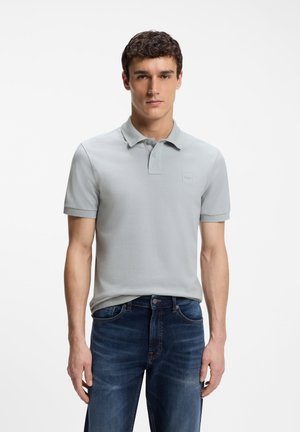 Άνδρας που φοράει ανοιχτό γκρι πόλο και σκούρο μπλε τζιν, στέκεται μπροστά σε λευκό φόντο, κοιτώντας μπροστά με ουδέτερη έκφραση.